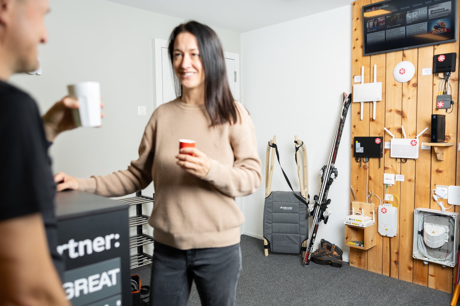 Zwei Personen unscharf im Vordergrund am Kaffee trinken und im Hintergrund Skis und einen Schlitten sowie Internet Router. Dominik Taeuber.